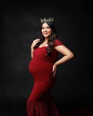 An elegant maternity portrait in a fitted red gown with crown against black backdrop.