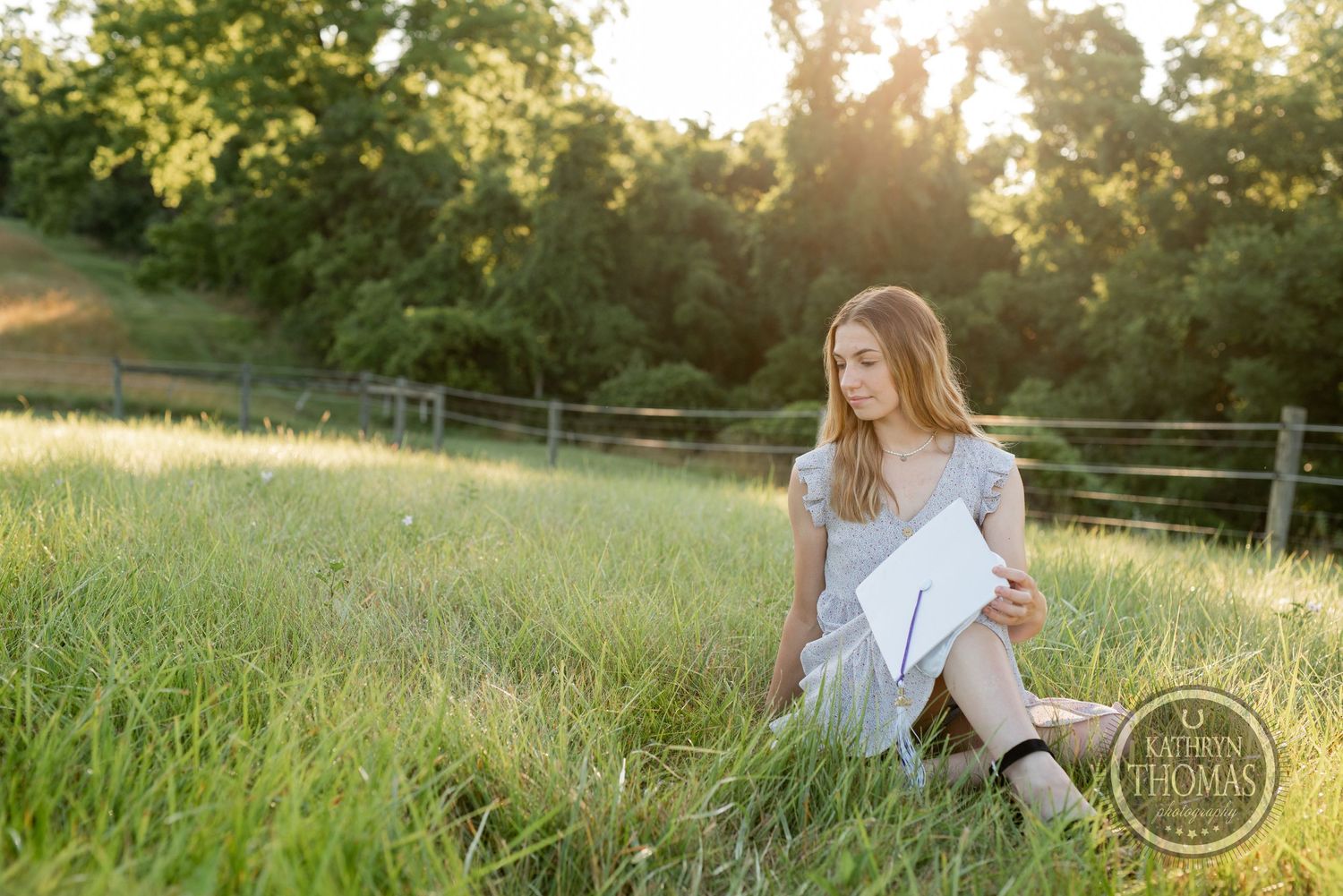 Graduate in flowy dress sits among tall grass during sunset graduation photoshoot.