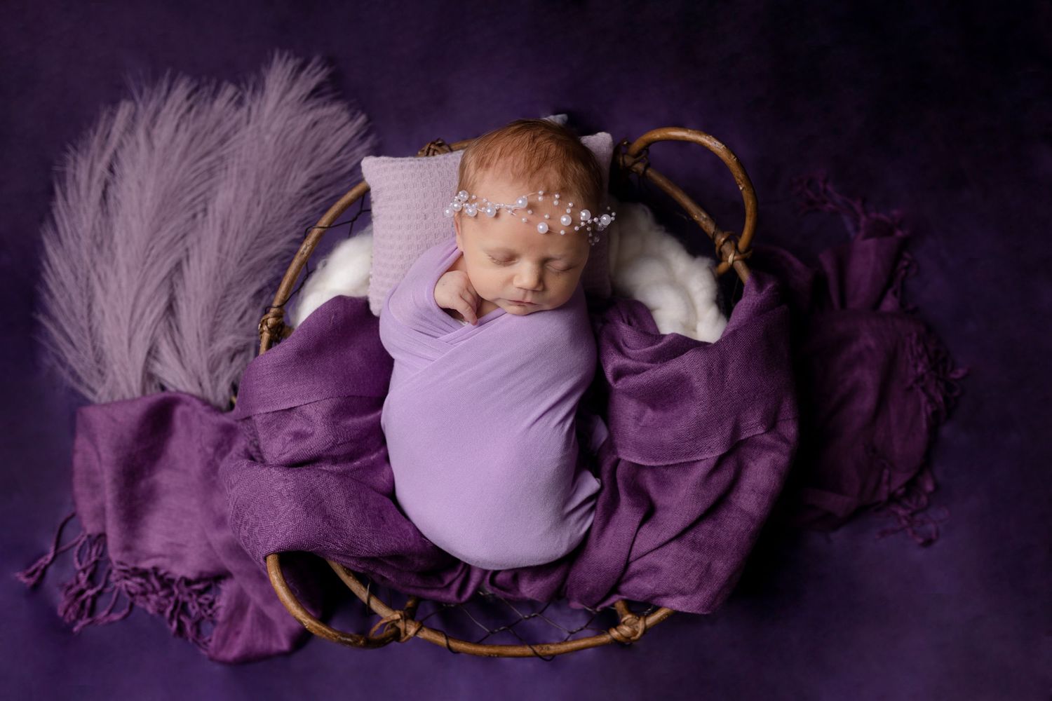 Newborn baby wrapped in purple swaddle with feather decorations on dark backdrop.