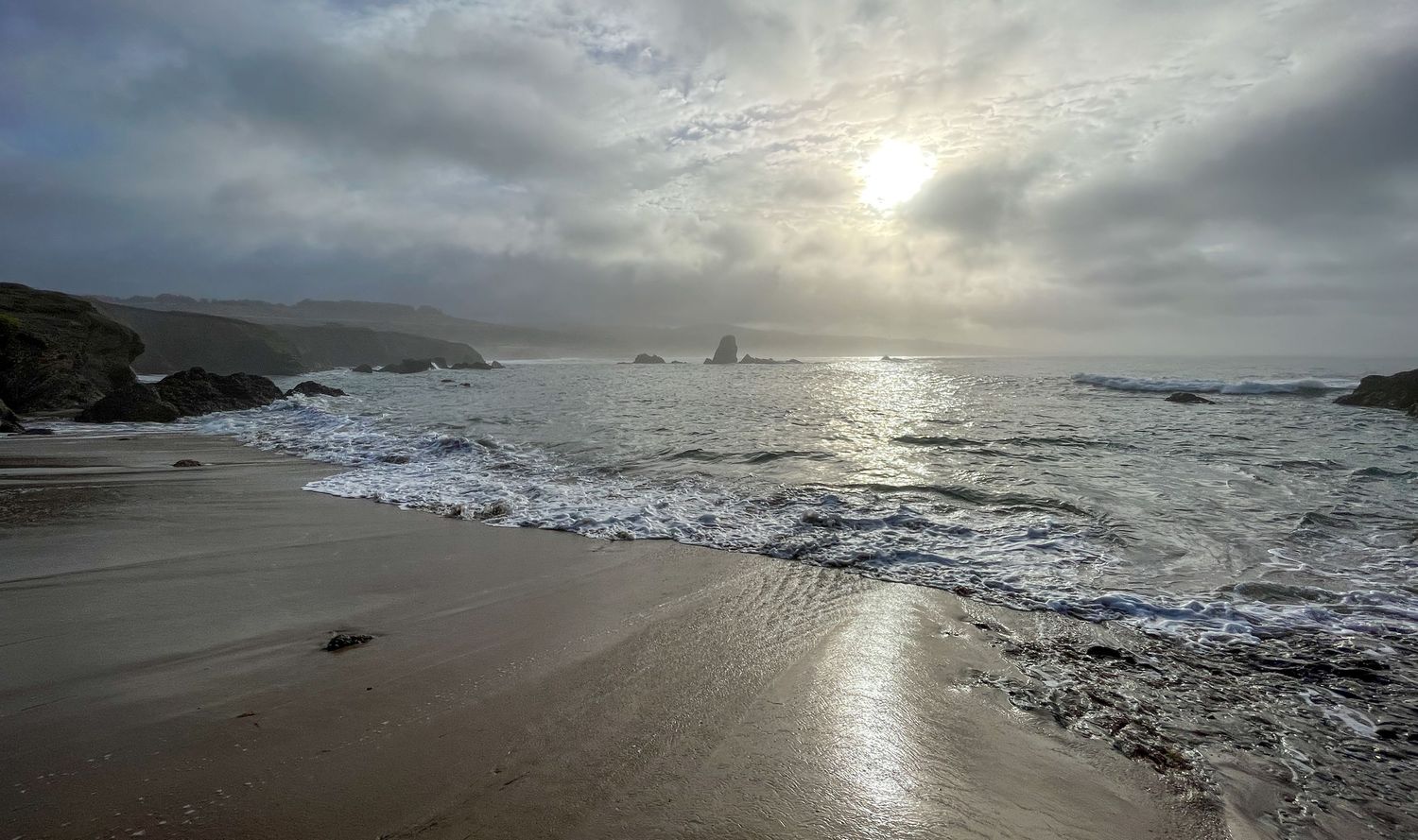 Rocky beach with waves gently lapping the shore, mist-covered cliffs, and a cloudy sky with the sun breaking through.