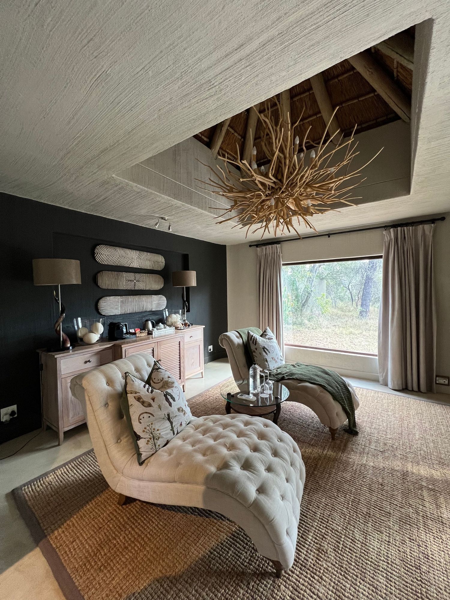 Modern safari lodge bedroom with unique twig chandelier and elegant chaise lounges facing panoramic window.