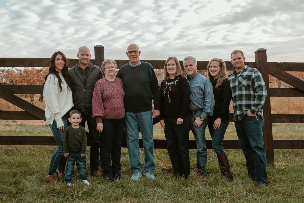 Large family gathering posing for a panoramic photo against wooden fence in rural setting.