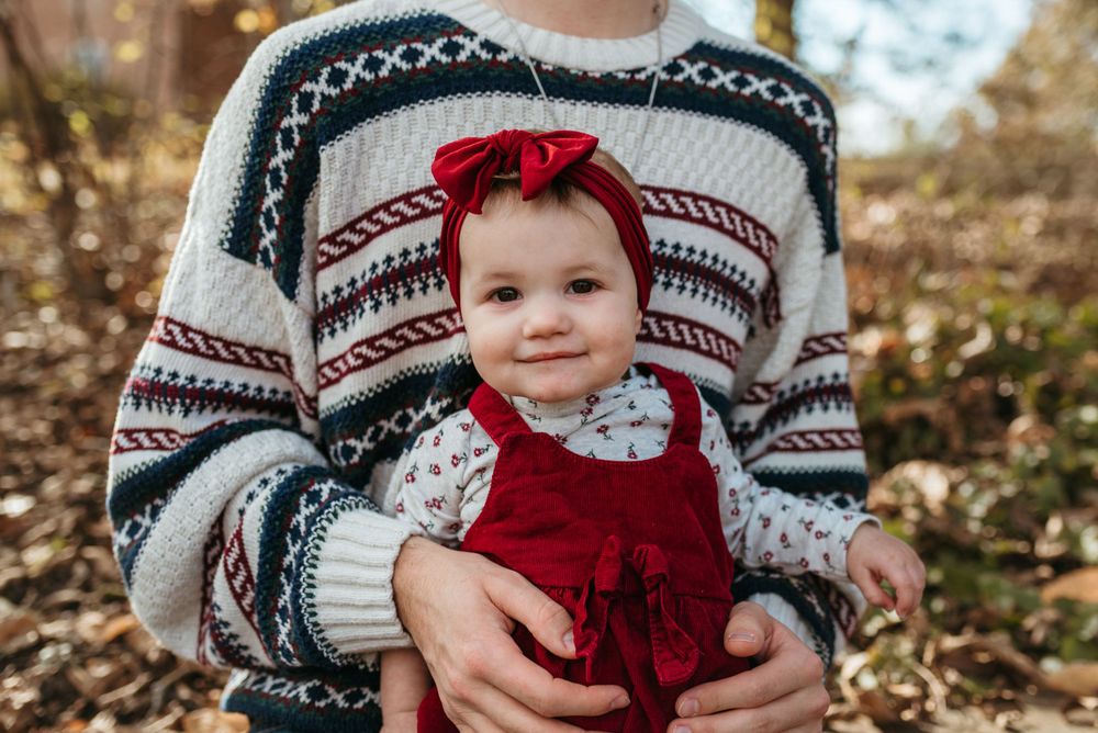 A baby in a red dress and bow sits against a festive winter sweater background.