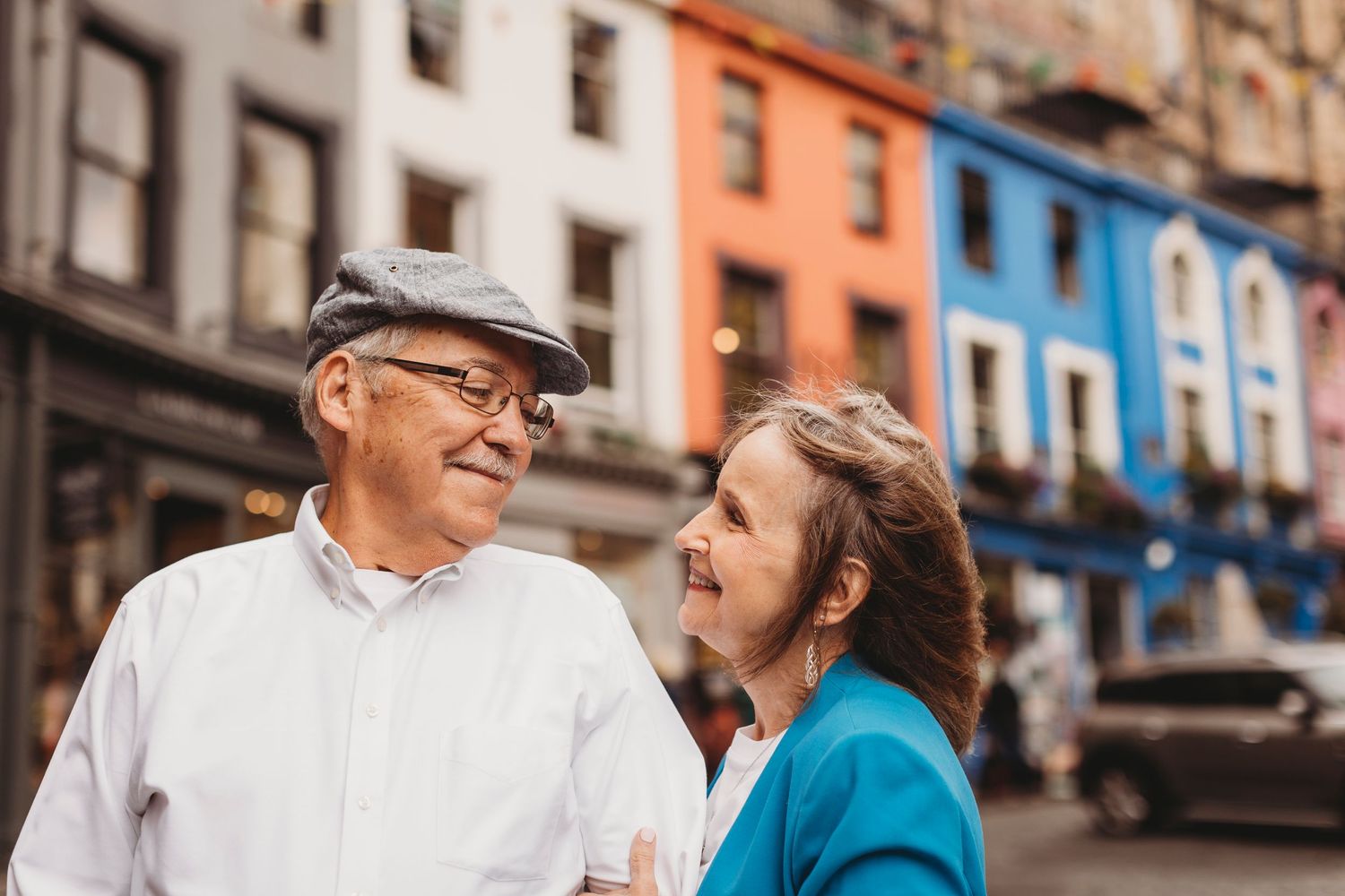 An elderly couple shares a tender moment on a city street with colorful buildings in Edinburgh.
