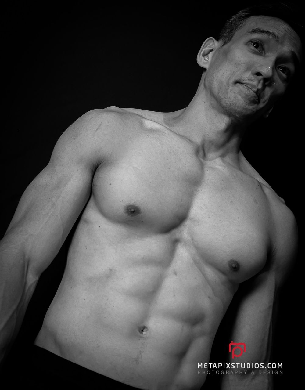 Black and white photo of a fit shirtless man with defined abs posing against dark background.