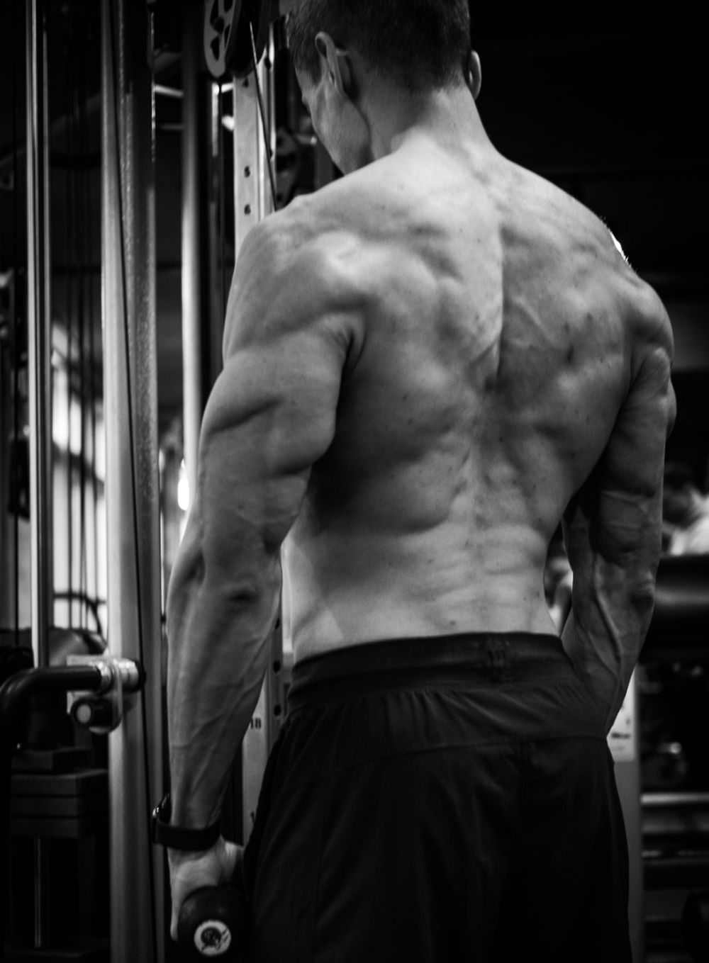 Black and white photo of a muscular man's back showing defined muscles at a gym cable machine.