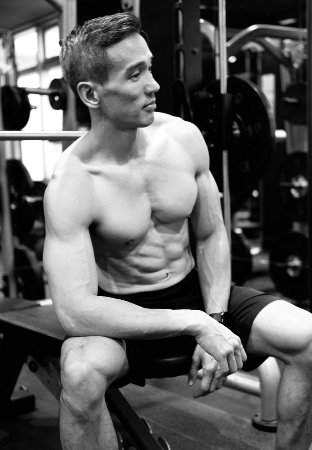 Black and white photo of a fit shirtless man sitting on a gym bench showing defined muscles.