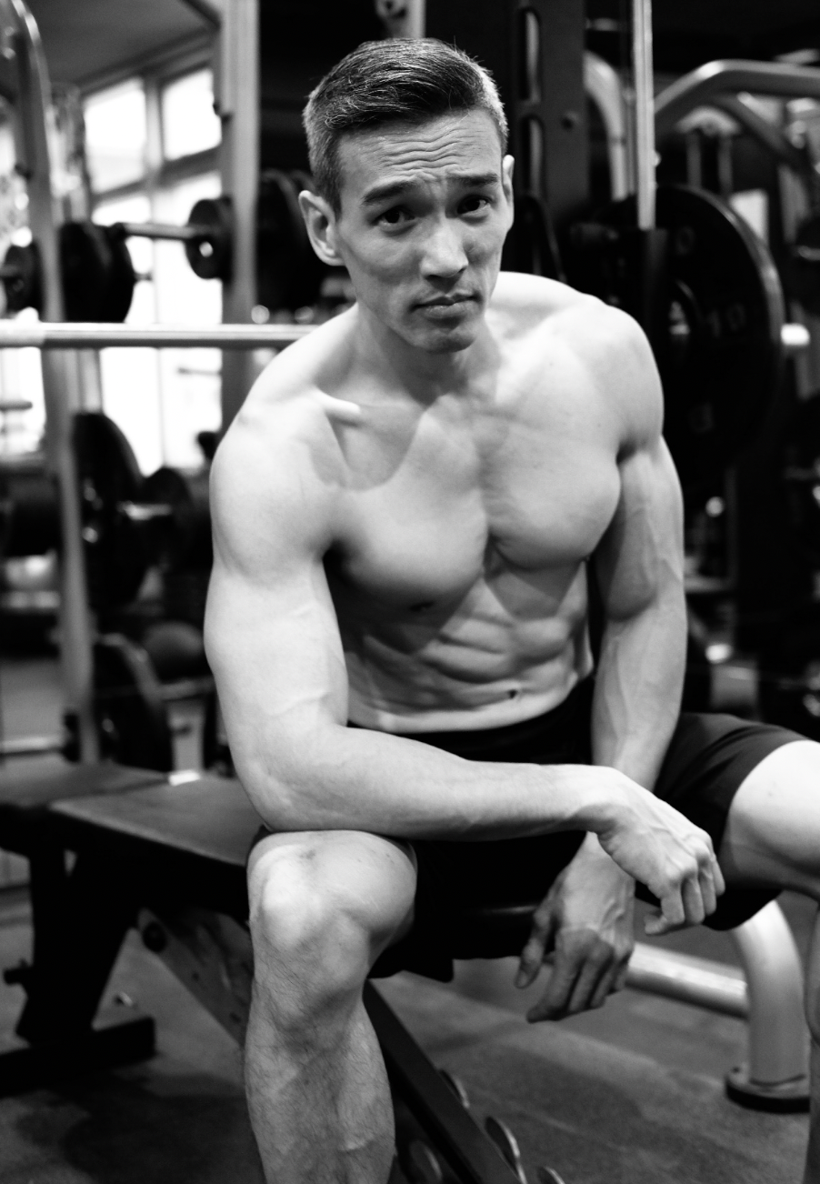 Black and white photo of a lean muscular shirtless man sitting on a gym bench posing.