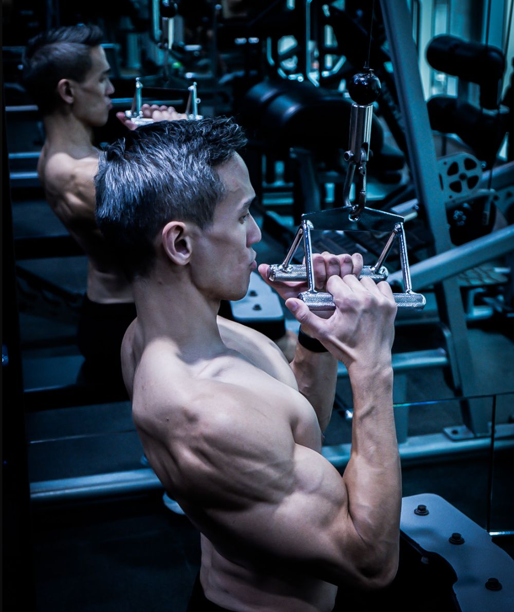 Muscular shirtless man pulling a cable machine attachment in a gym, reflected in a mirror.