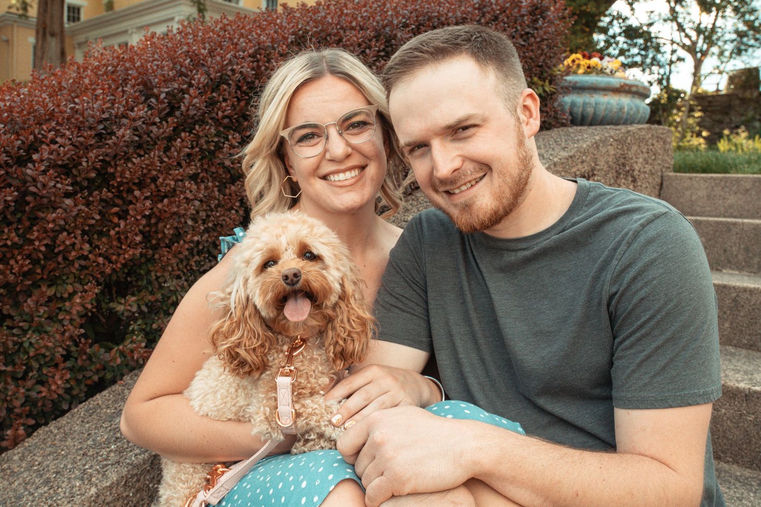 A couple poses outdoors with their goldendoodle puppy against red foliage.