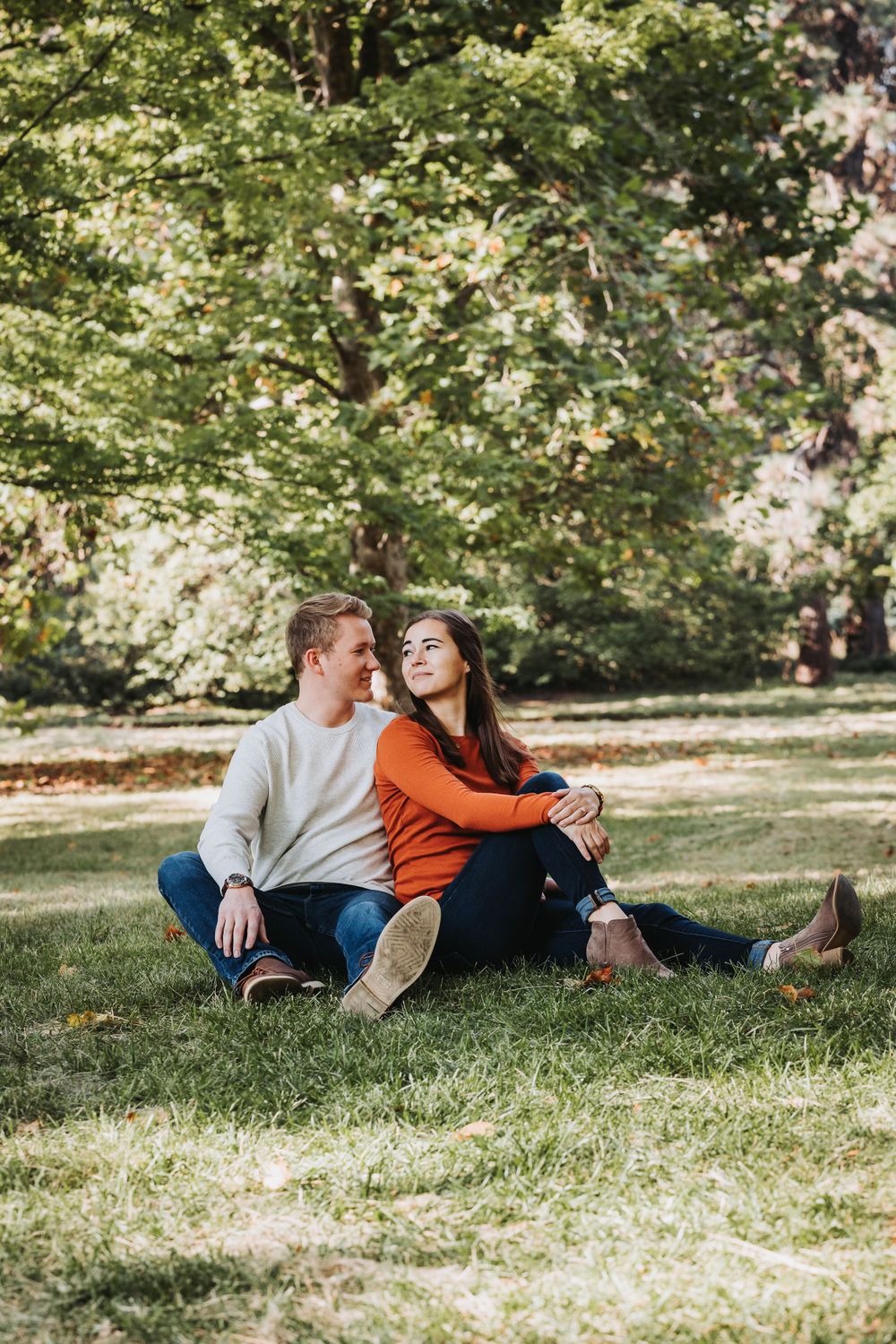 A couple enjoying their engagement session, sitting together on the grass in a sunny park, surrounded by nature in Finch Arboretum