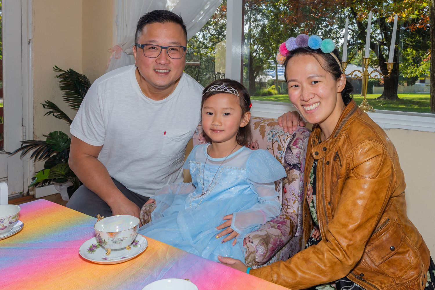 Asian family posing at rainbow tablecloth tea party table with girl in Cinderella costume.
