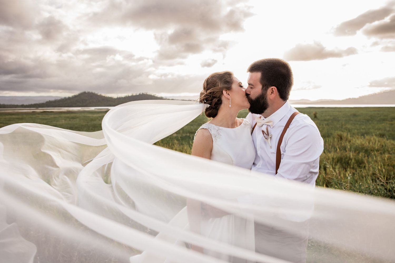 Wedding couple embraces in field at sunset as bride's veil dramatically flows in the wind.