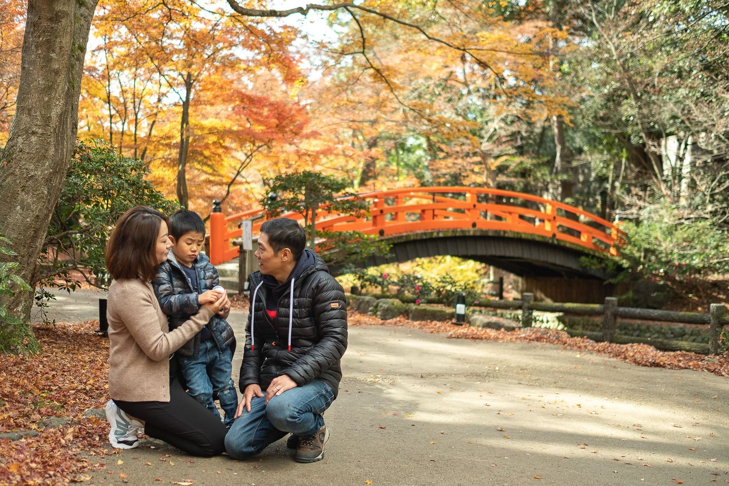 Family sitting together on a path in a Japanese garden with stunning autumn foliage and red bridge.