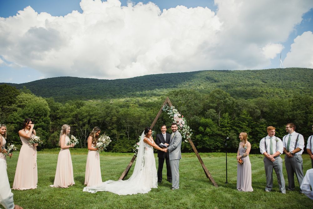 Wedding ceremony in progress against green mountain landscape with rustic wooden triangle arch.