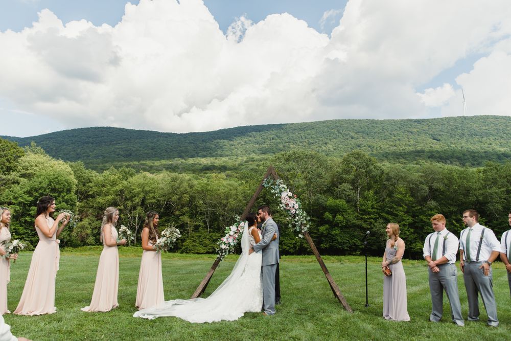 Outdoor wedding ceremony with mountain backdrop featuring wedding party gathered around wooden triangle arch.