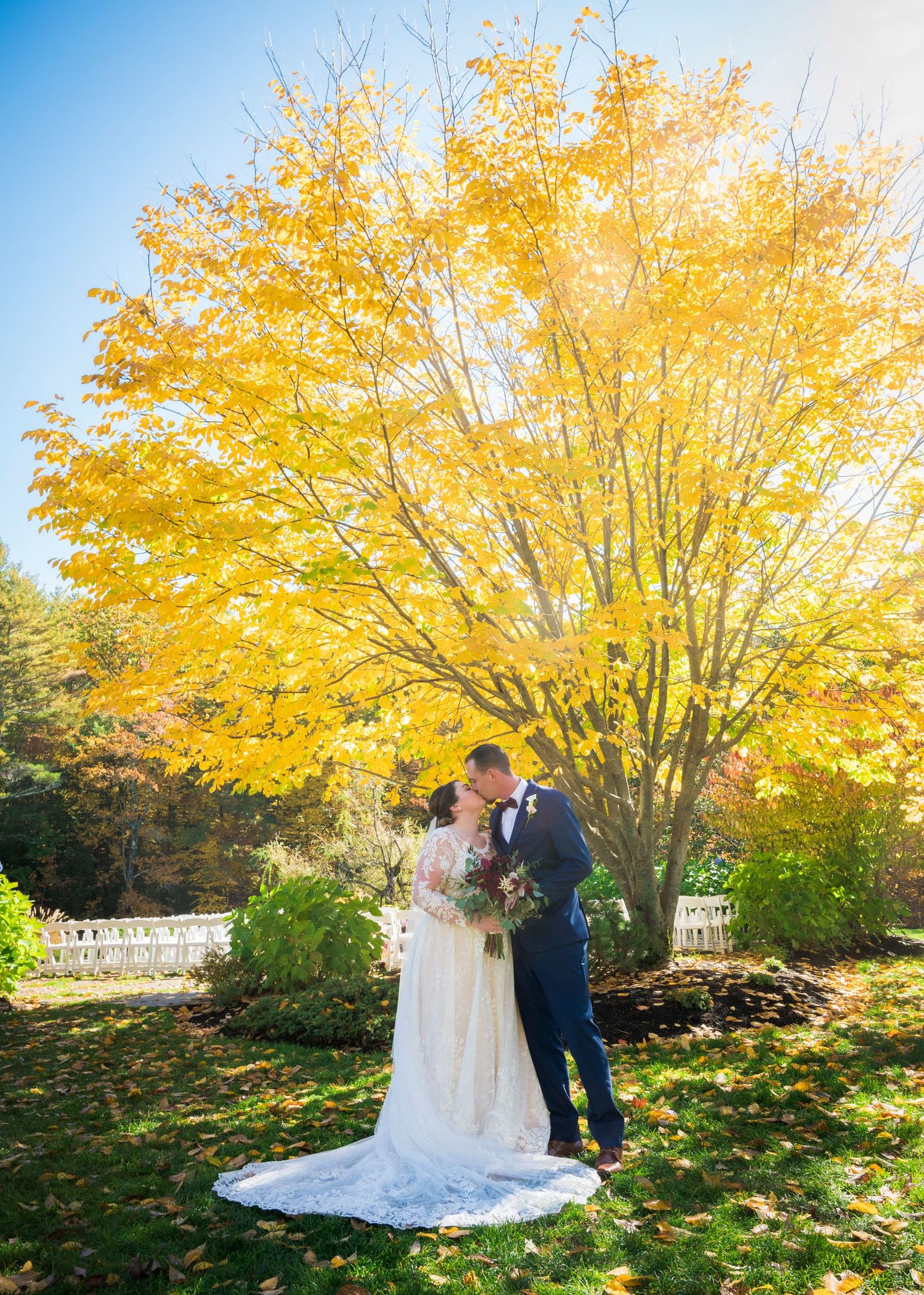 A couple stands beneath a vibrant yellow autumn tree in formal wedding attire.