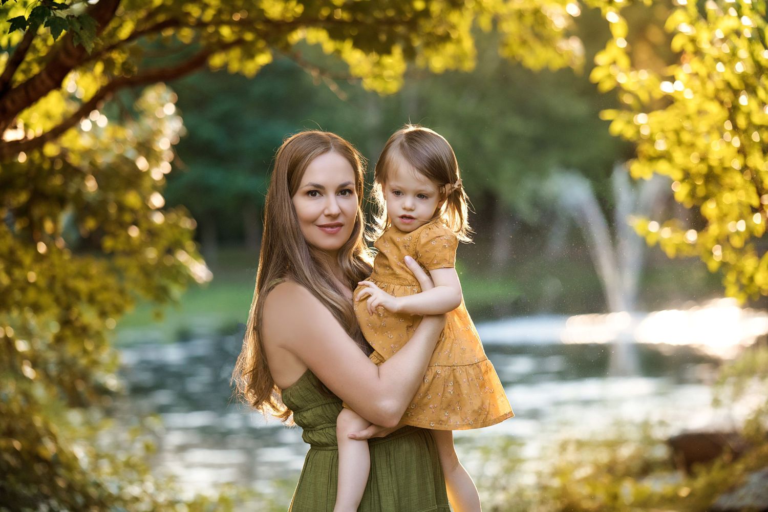 A parent and child share a tender moment amid golden sunlit trees near a pond.