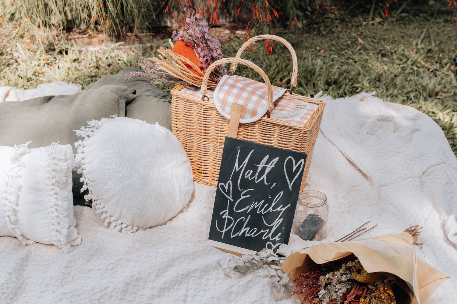 A rustic picnic setup with wicker basket, white blanket, and chalkboard sign for a proposal.