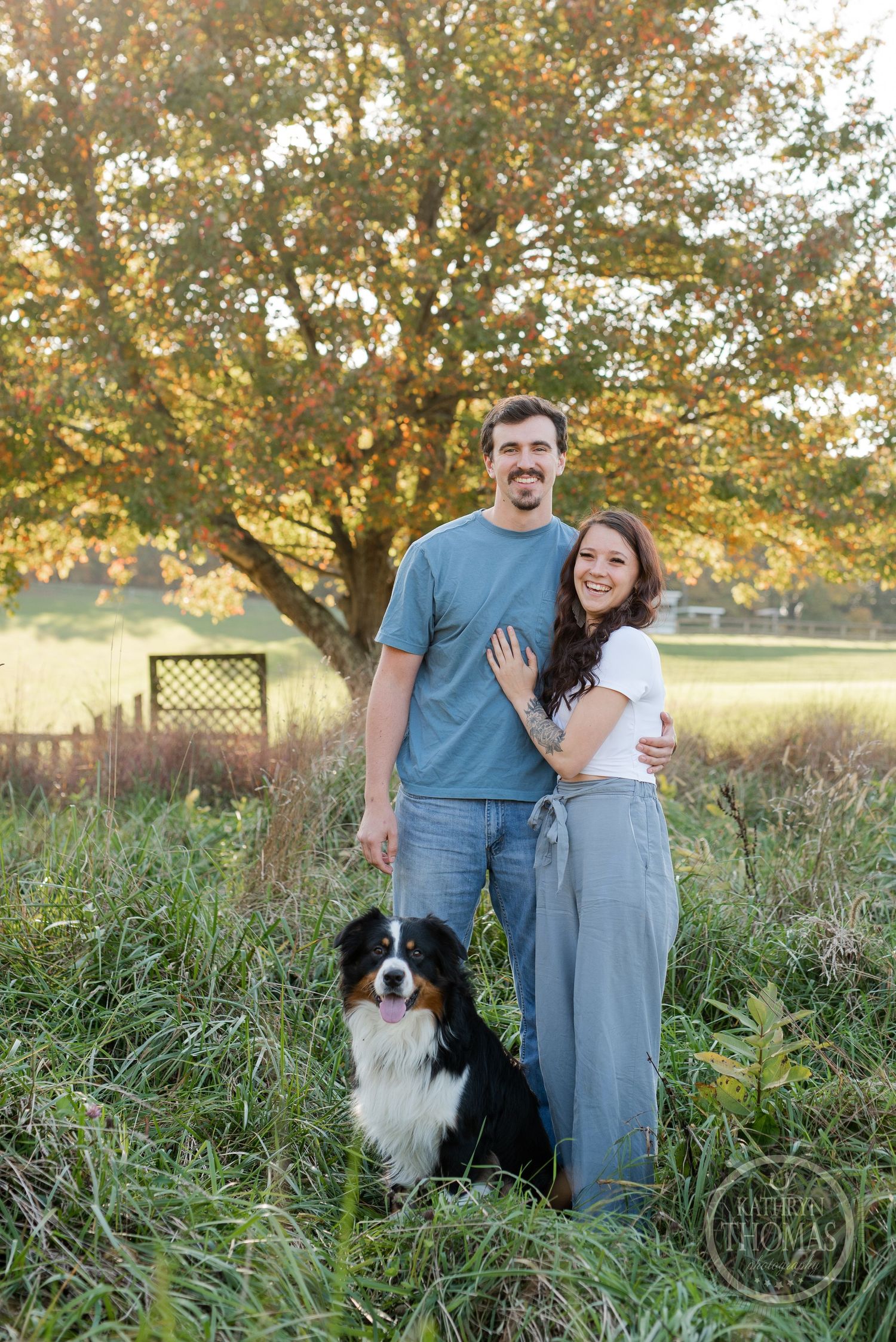 A couple poses with their Border Collie in a rustic outdoor setting during golden hour.