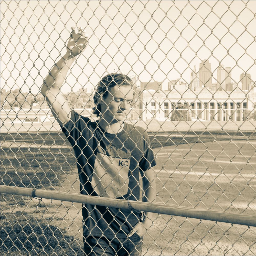 Vintage-style photo of person gesturing through chain-link fence in urban setting.