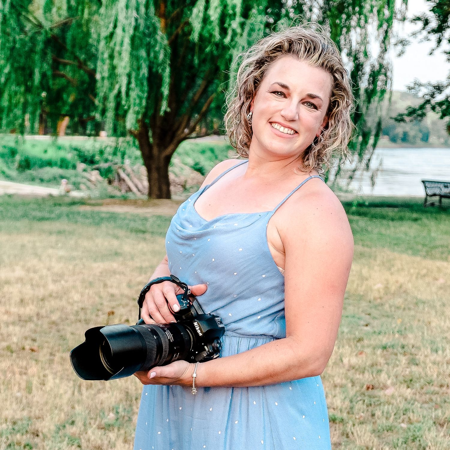 Stephanie Coulter, photographer, in blue dress holding camera outdoors under willow tree by Ericka Faust