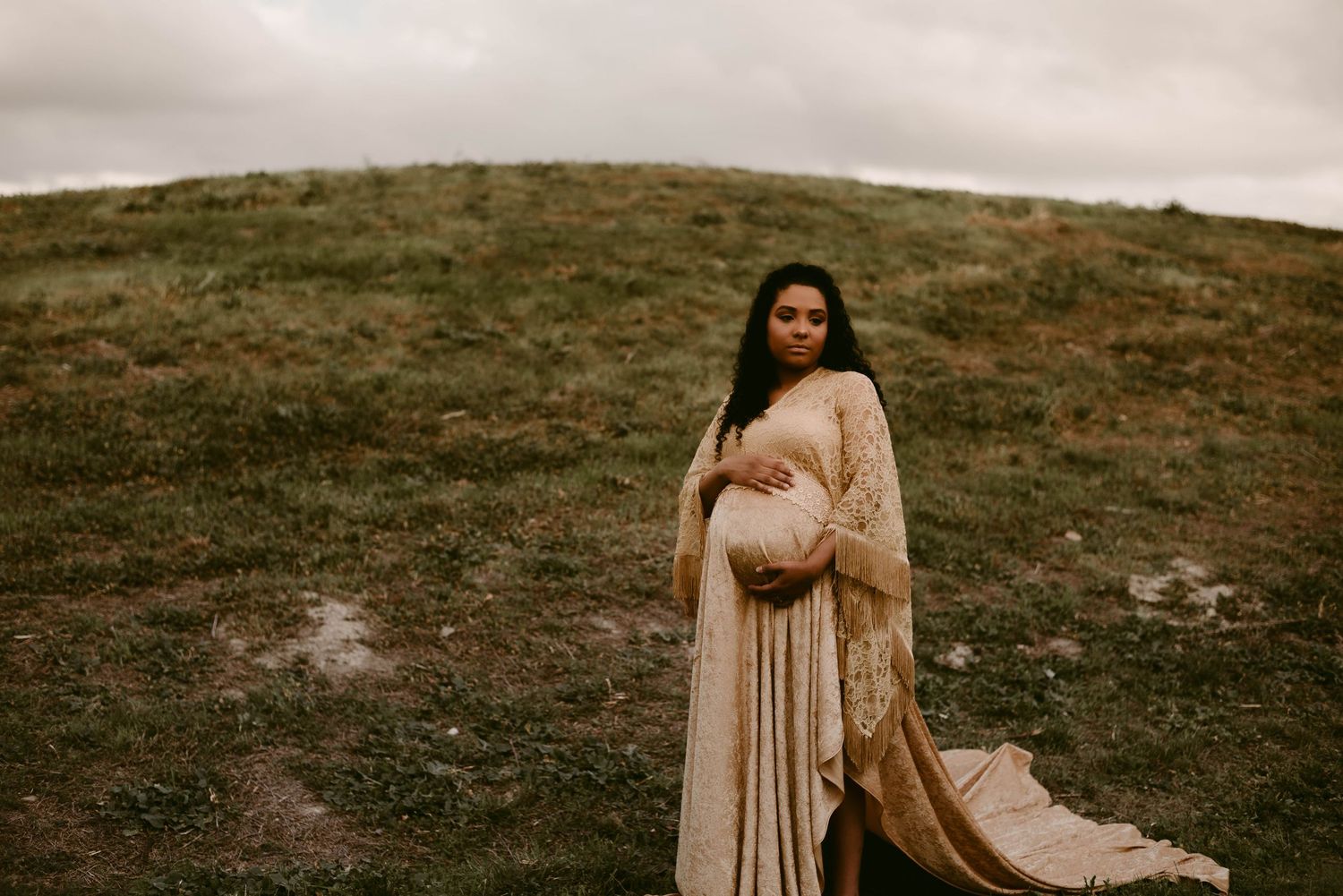 Moody maternity portrait on hillside with flowing champagne dress and dramatic sky.