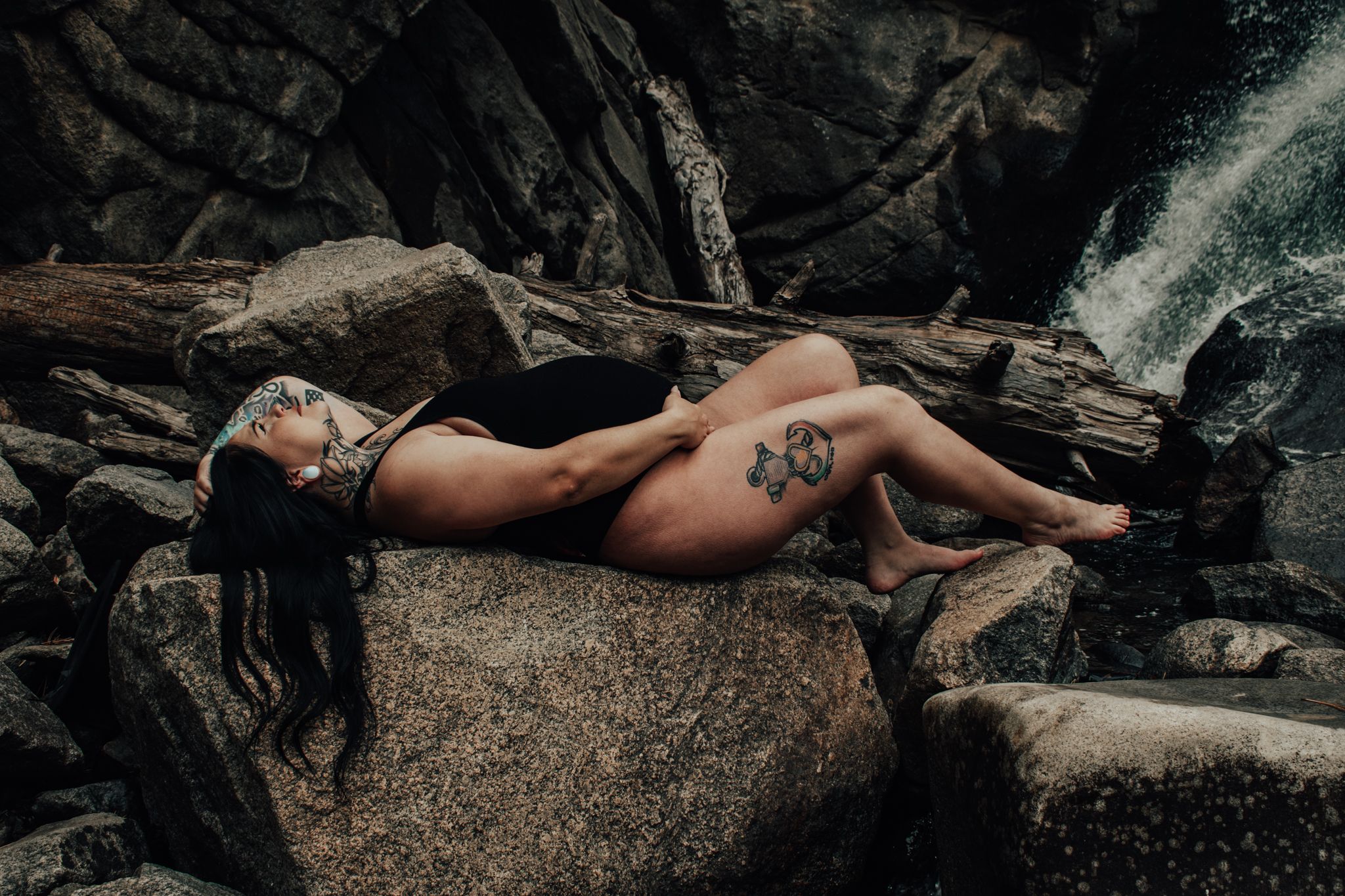 Tattooed woman reclines on rocky boulders near a waterfall in a nature maternity shoot.