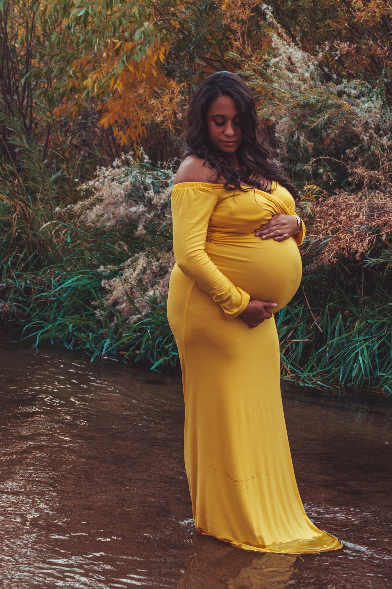 Pregnant woman in yellow off-shoulder gown standing in a stream surrounded by autumn foliage.