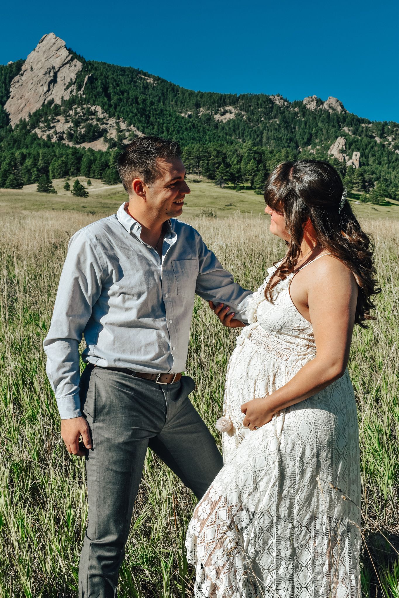 Couple shares a tender moment in green meadow with rocky Flatirons mountains in background.