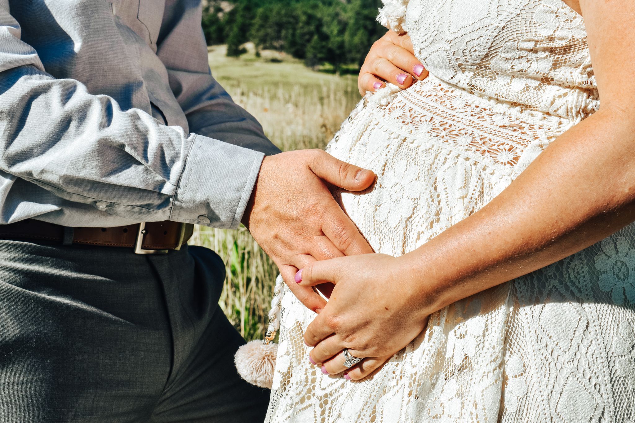 Close-up of couple holding pregnant belly in lace dress outdoors in sunny field.