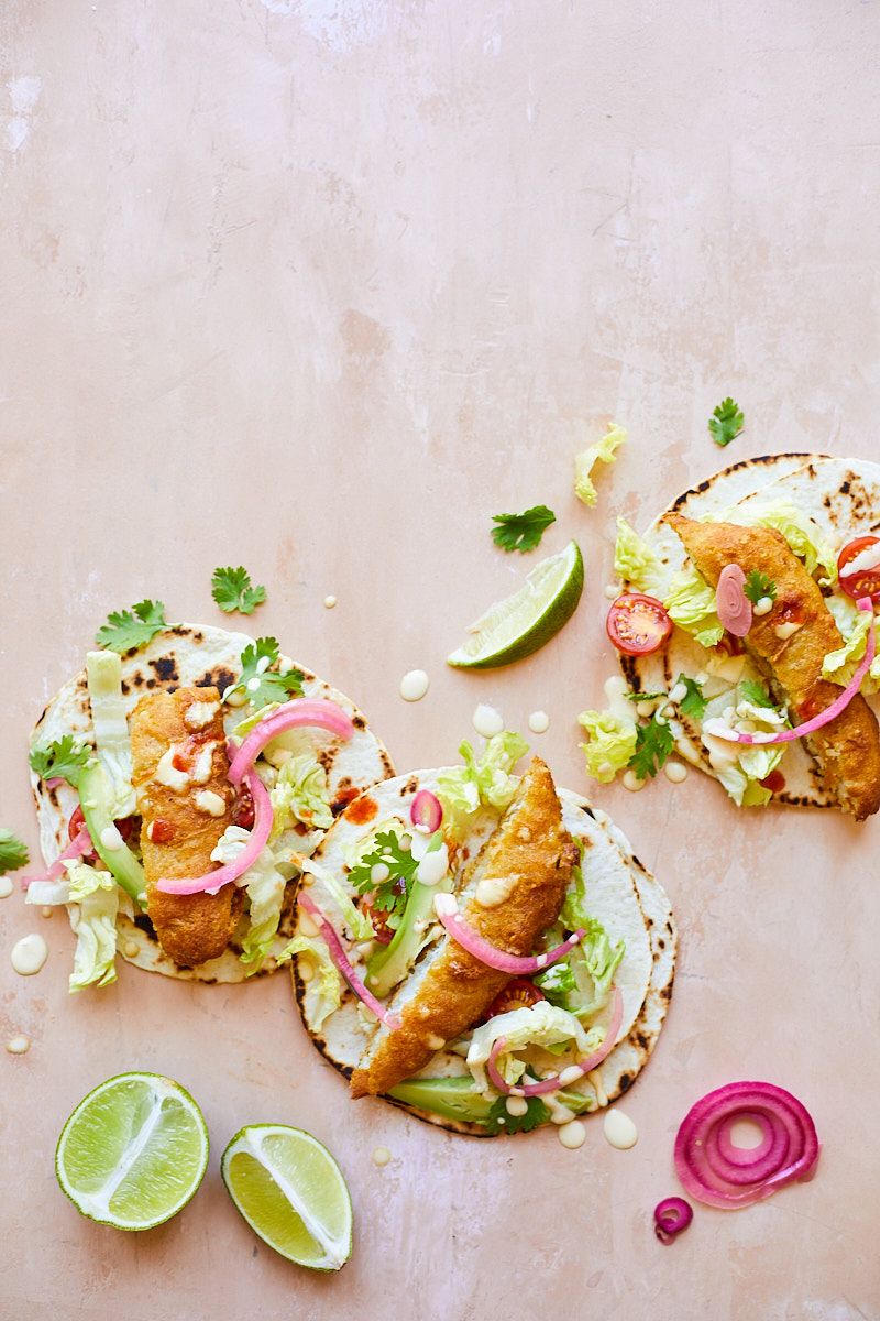 Overhead shot of fish tacos with lime wedges and pickled onions on a pink surface.