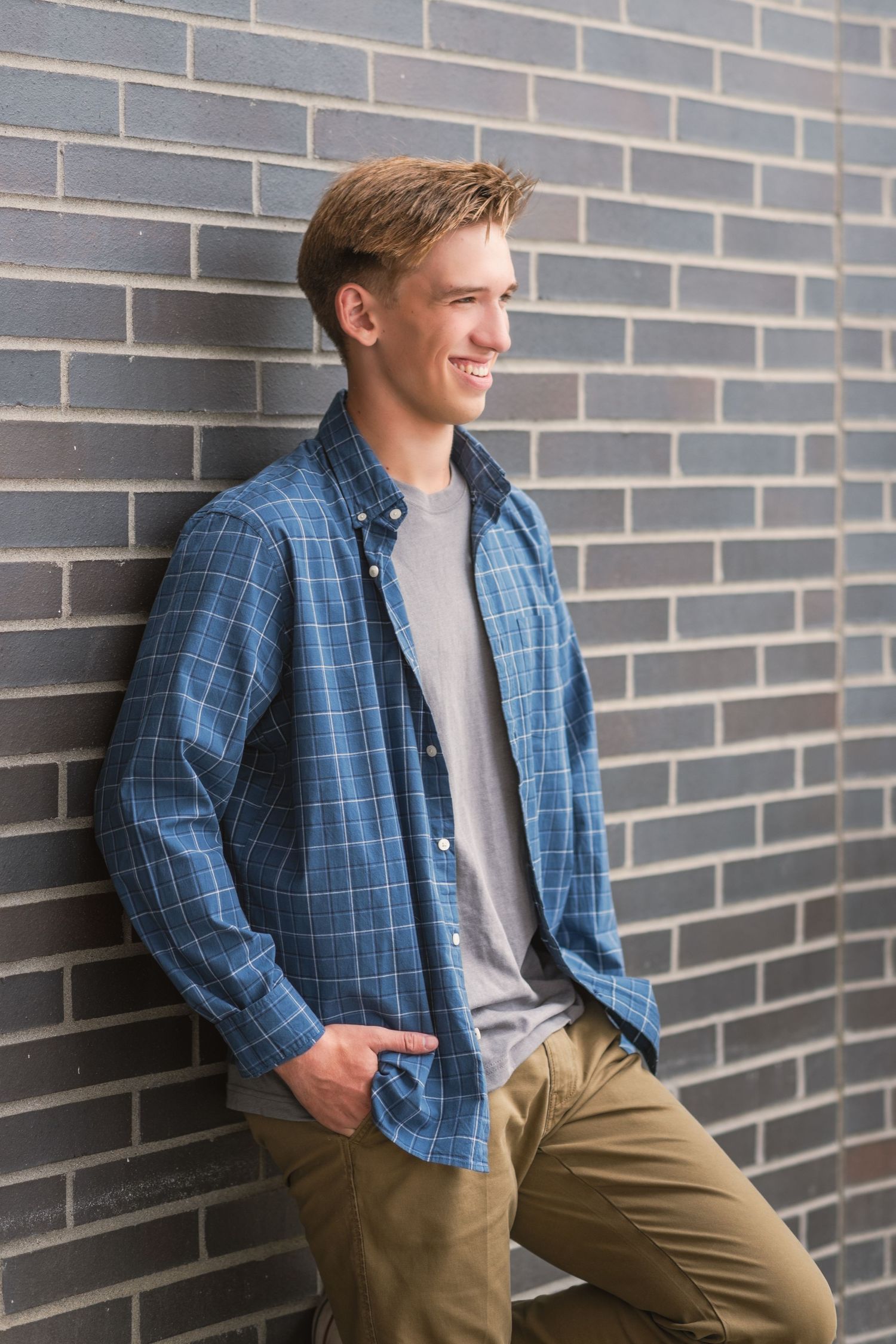 senior from lakeridge highschool leaning against dark brick wall with hands in his pockets looking away from camera