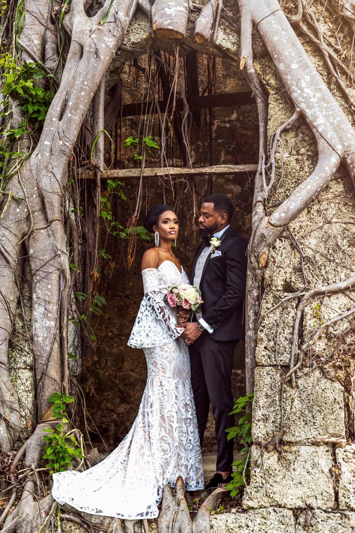 A wedding couple poses in an archway framed by tree roots and vines at a historic stone building.