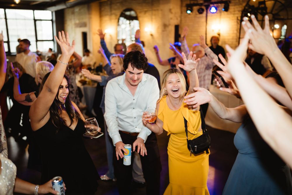 Wedding guests dance energetically with raised arms on a dark dance floor at a reception celebration.