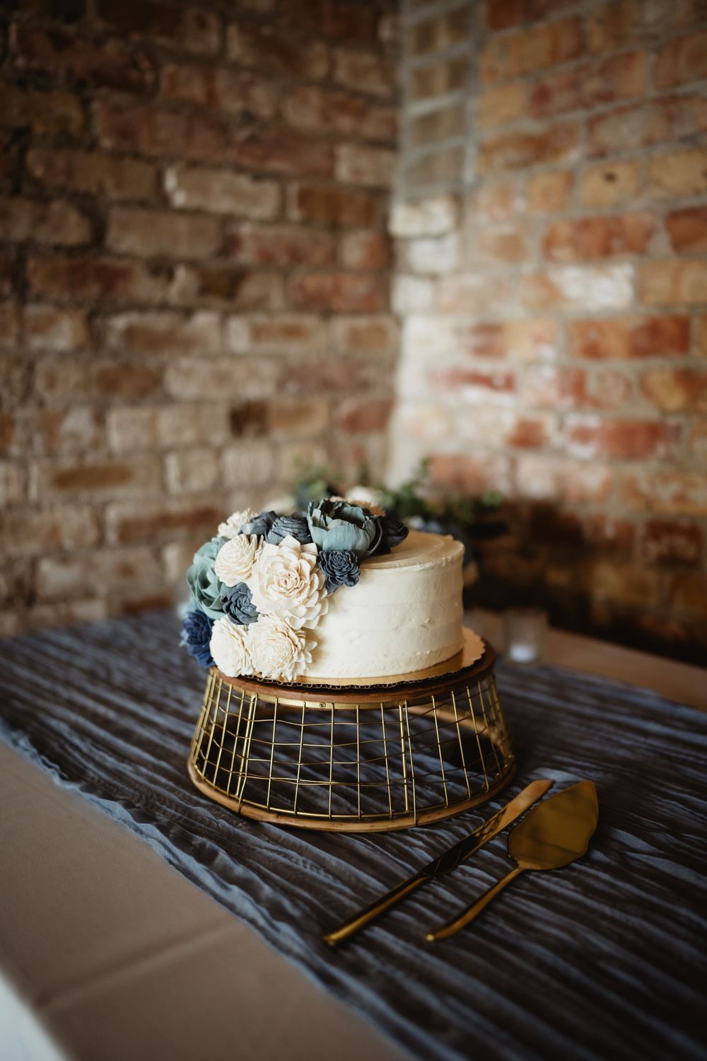 Elegant white wedding cake with blue and cream flowers on gold stand against brick wall.