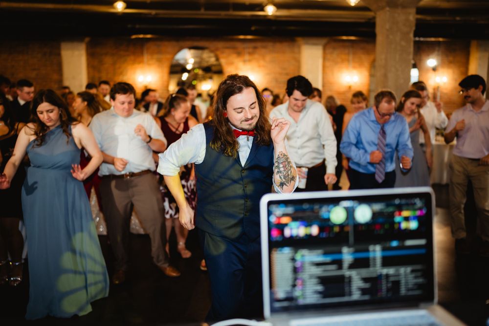 A DJ performs at a wedding reception with guests dancing in the background and a laptop visible in the foreground.