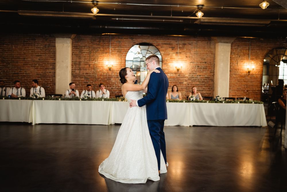 A couple shares their first dance in a rustic brick-walled reception venue with elegant lighting and white-clothed tables.