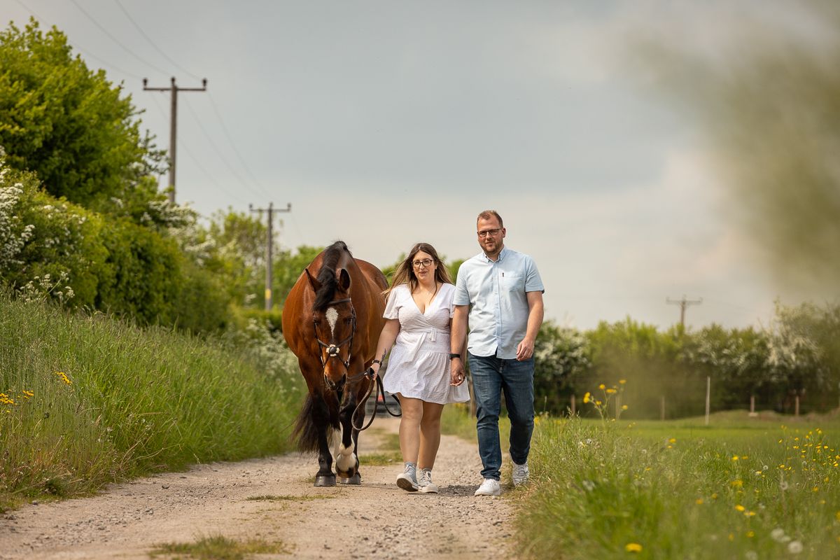A couple walking their horse down a country path surrounded by lush green grass and wildflowers.