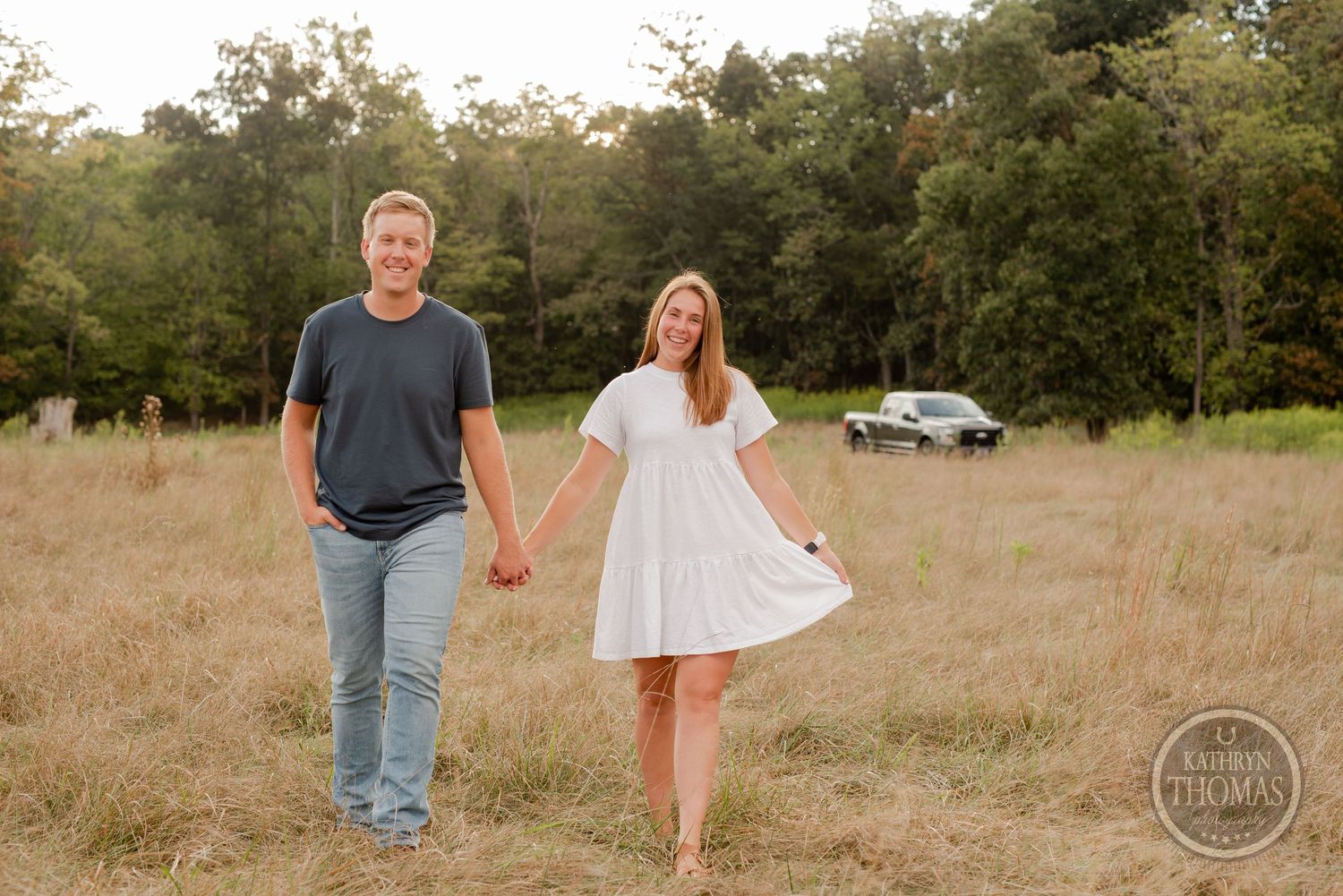 Couple walking hand in hand through rustic field during sunset photo shoot.