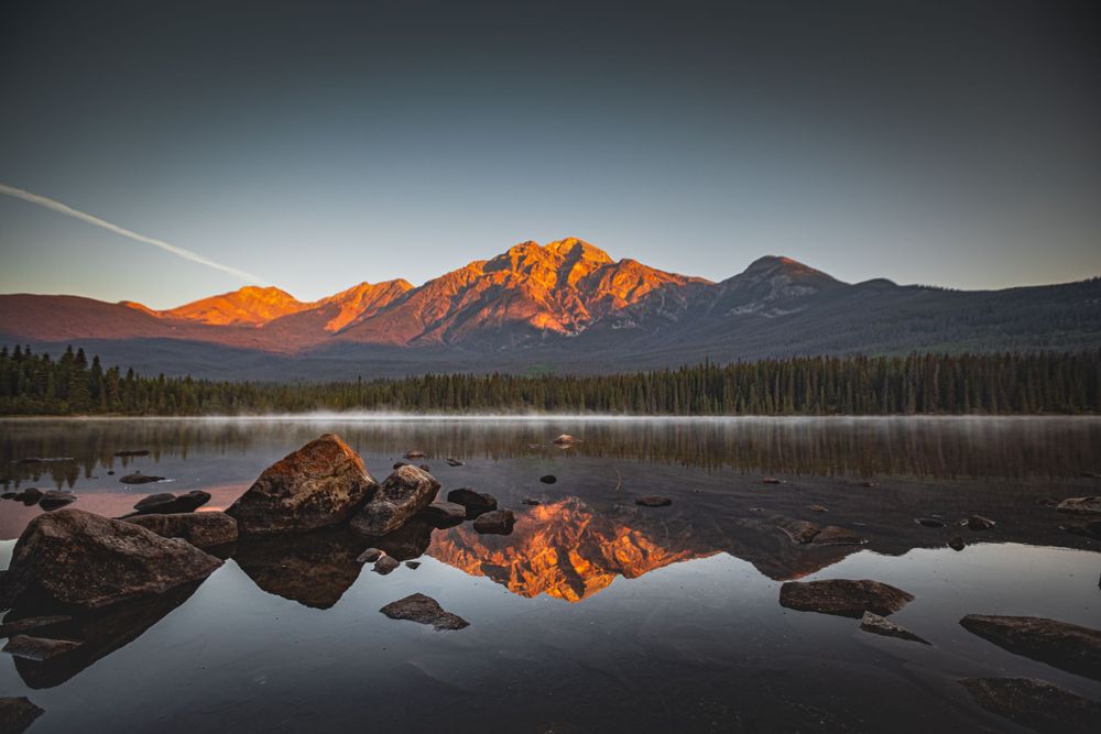Snow-capped mountain peak glows orange at sunrise, perfectly mirrored in still mountain lake.