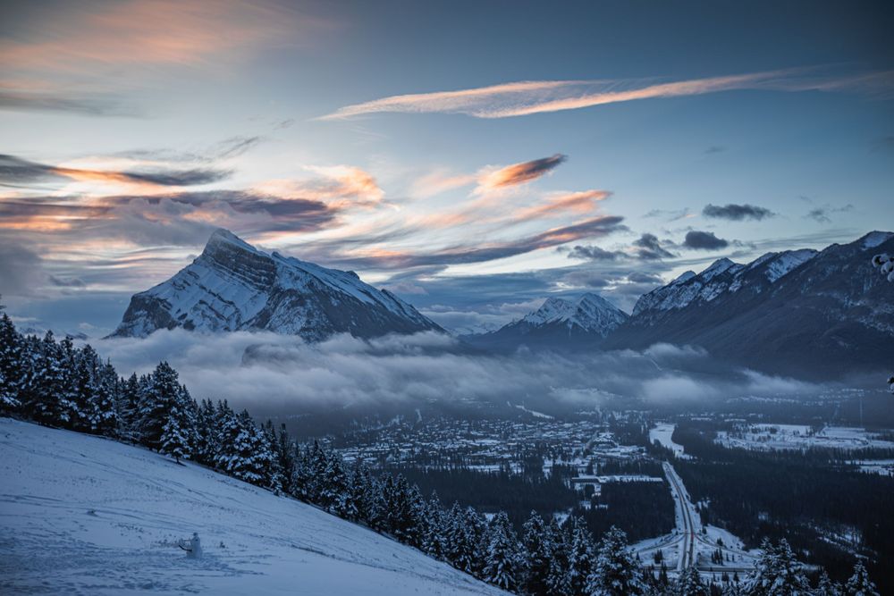 Snowy mountain landscape at dusk with dramatic clouds and a valley town nestled below in winter.