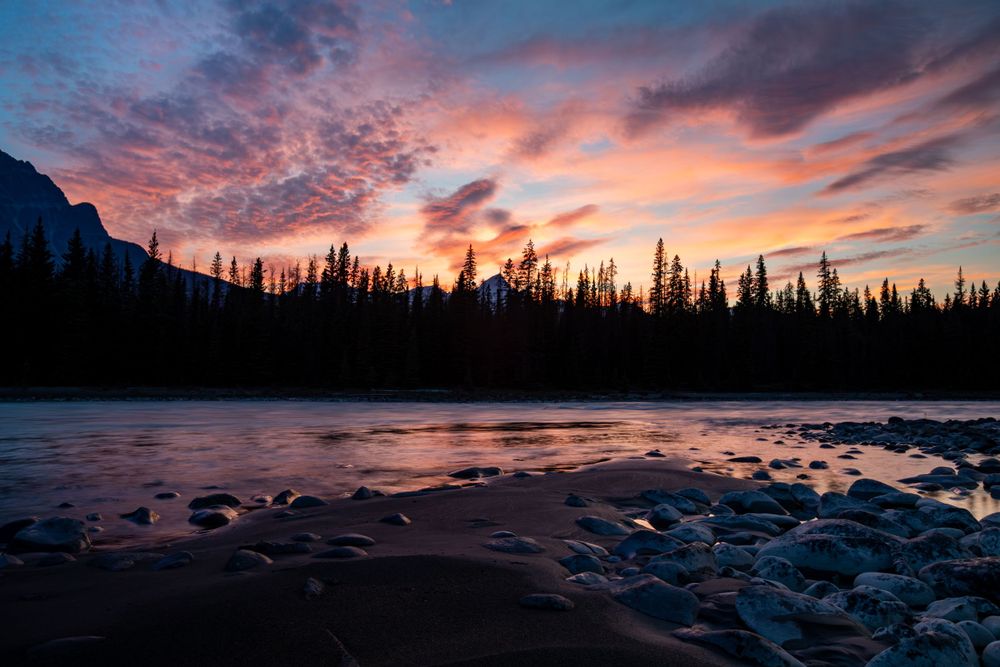Vibrant sunset sky reflects in rocky mountain lake with silhouetted pine forest.