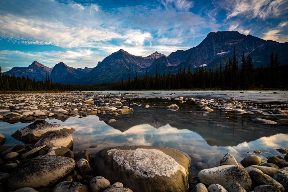 Rocky riverbed reflects mountain peaks under dramatic clouds in scenic mountain landscape.
