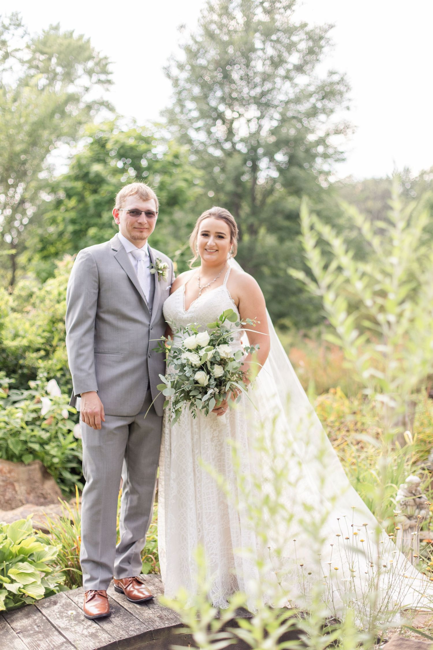 Wedding couple poses in a lush garden setting with white floral bouquet.