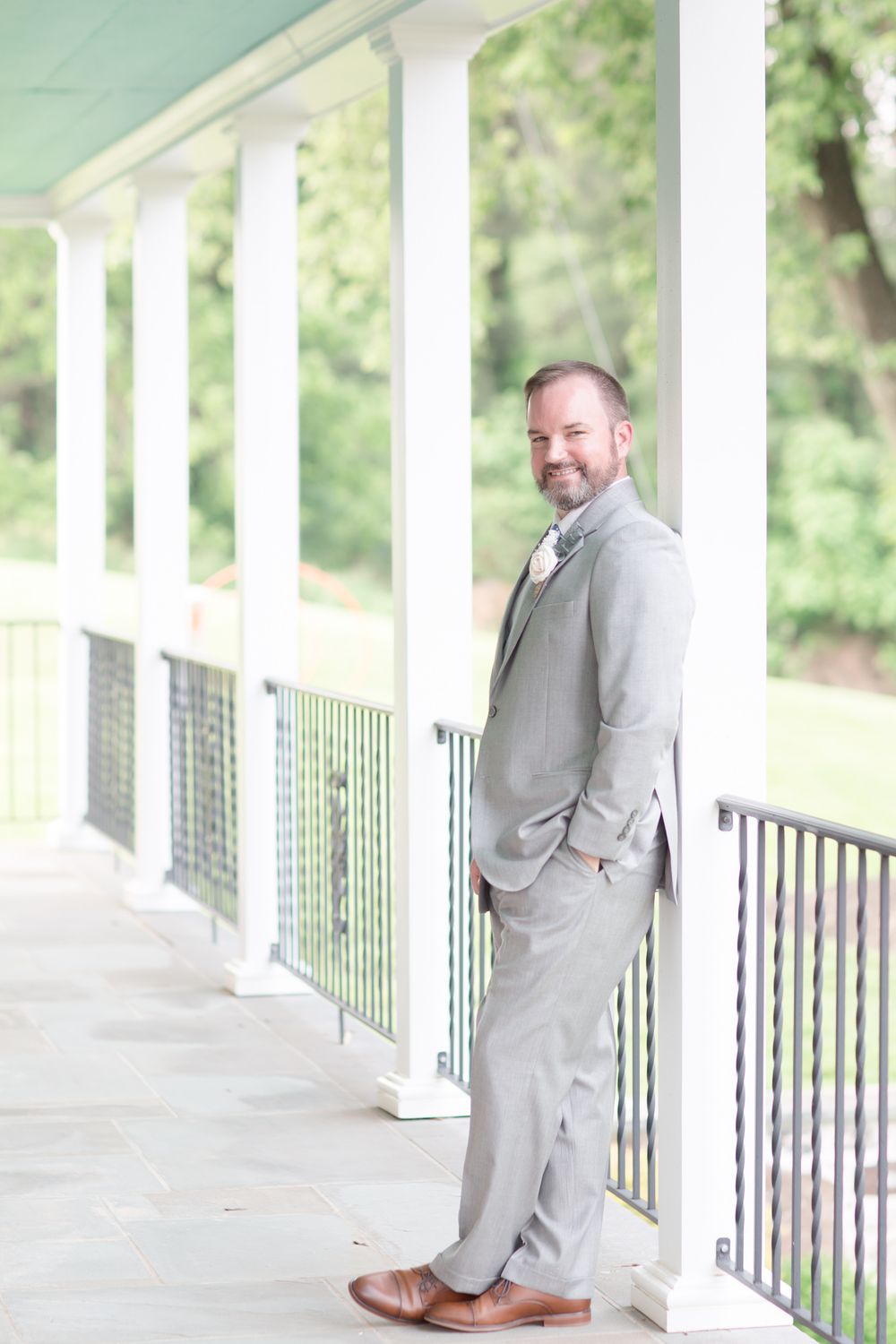 Elegant groom in light gray suit stands on columned porch with greenery backdrop.