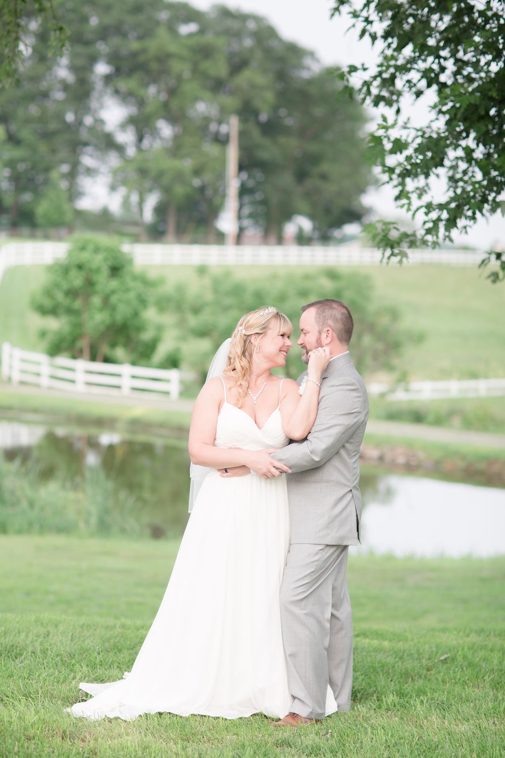 Soft, ethereal wedding portrait beside a pond with white fencing and lush greenery.