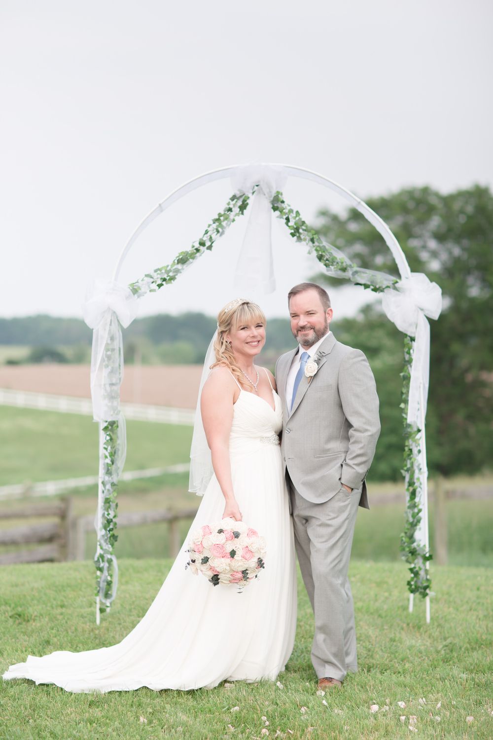 Wedding ceremony under a decorated white arch with greenery in a pastoral setting.