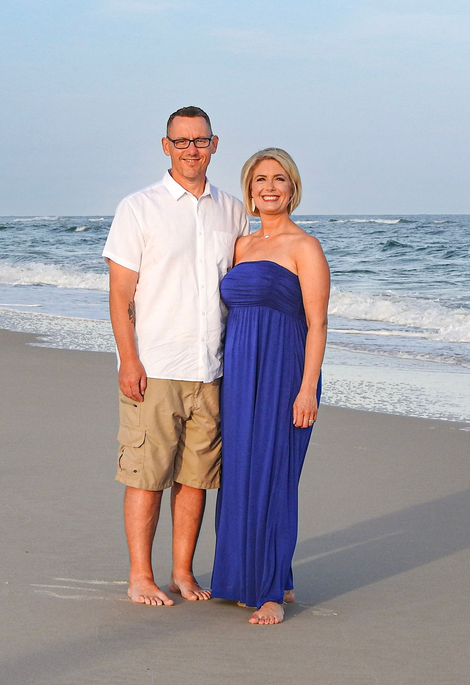 Casual beach portrait of couple standing together on sandy shore with ocean waves behind them.