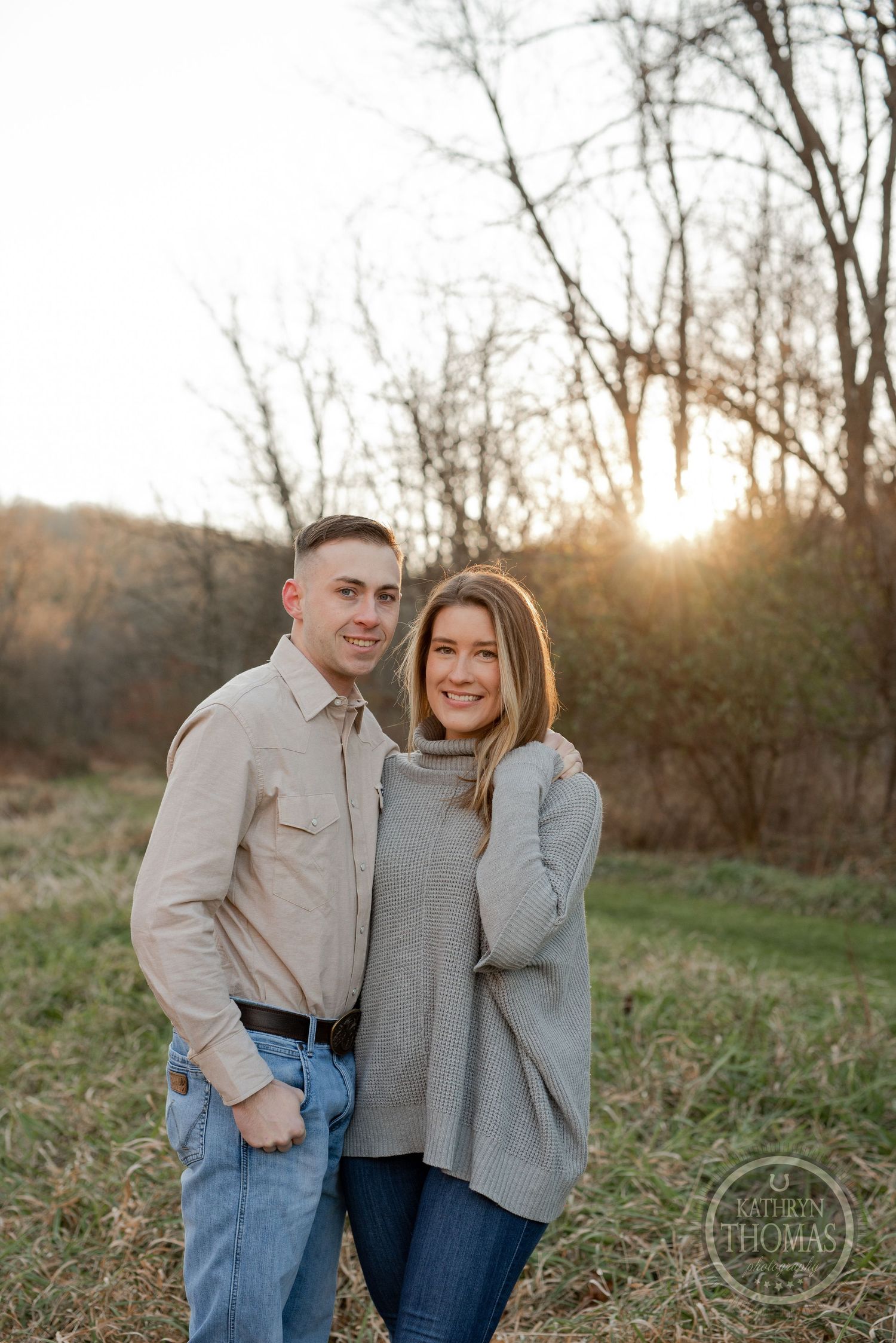 A couple poses together in casual autumn attire during golden hour with bare trees in the background.